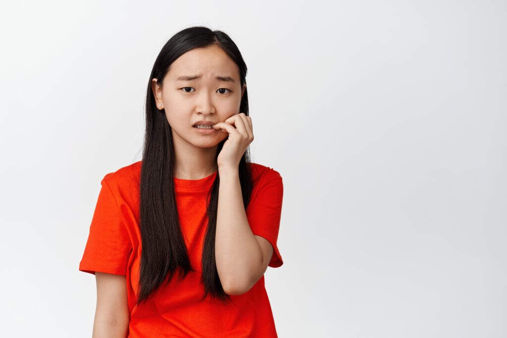 A young woman in a red t-shirt looking worried while biting her fingernail, exemplifying the habit of nail biting and its potential harm to dental health.