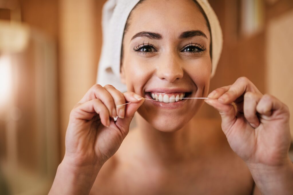 Smiling woman with a towel wrapped around her head, using dental floss to maintain oral hygiene.