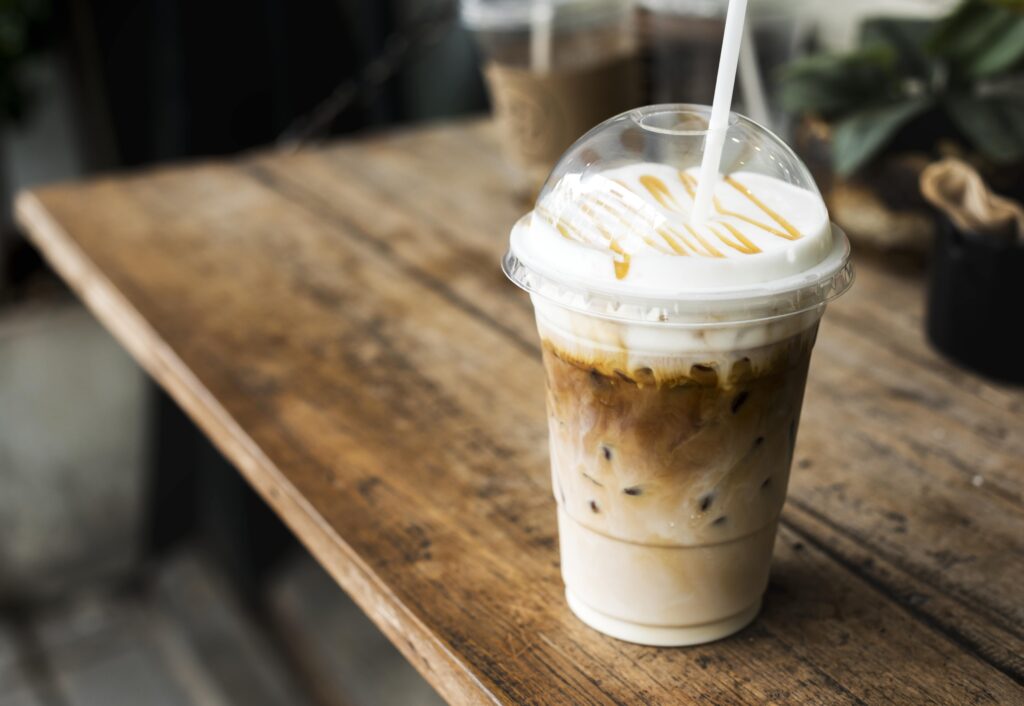 Iced caramel macchiato with whipped cream and caramel drizzle in a clear plastic cup with a straw, placed on a rustic wooden table, perfect for a summer refreshment or coffee break concept.