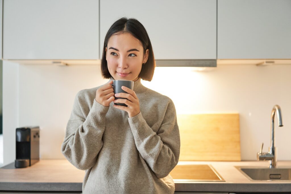 A cheerful Asian woman savoring her morning coffee in a well-lit modern kitchen, embodying a relaxed lifestyle compatible with clear aligner use
